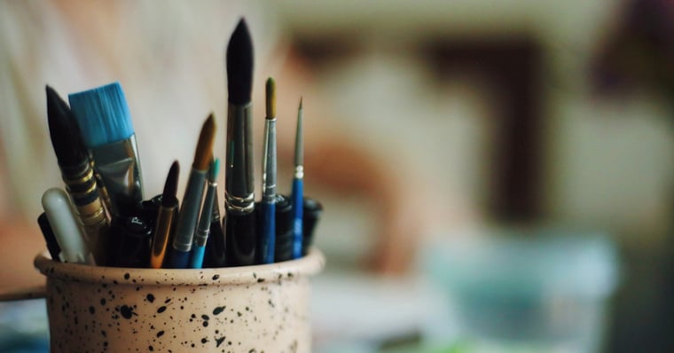 A collection of blue paintbrushes in a cup. In the background, a woman is working at a table.