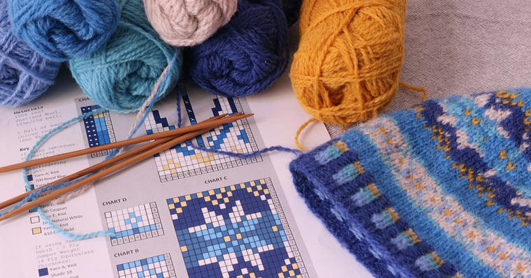 A knit hat next to knitting instructions, knitting needles, and balls of yarn.